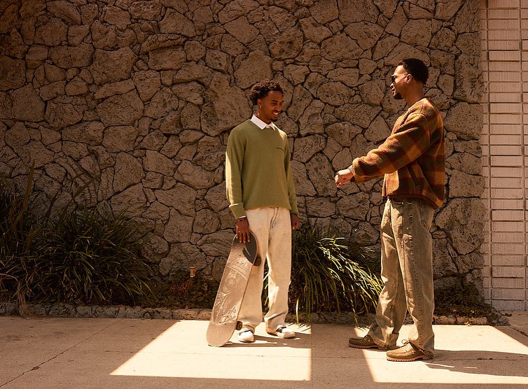 Two adult men in casual clothing stand outdoors on a sunny day, one holding a skateboard, with a stone wall and modern building behind them.