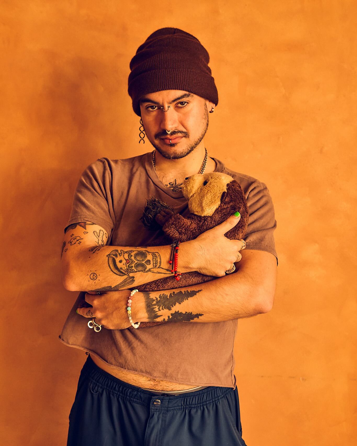 Adult male with tattoos wearing a beanie and cropped shirt, hugging a brown plush toy in a studio with an orange backdrop.