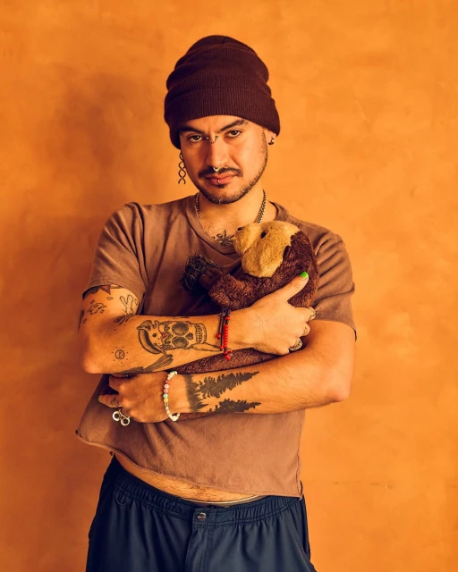 Adult male with tattoos wearing a beanie and cropped shirt, hugging a brown plush toy in a studio with an orange backdrop.