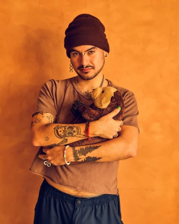Adult male with tattoos wearing a beanie and cropped shirt, hugging a brown plush toy in a studio with an orange backdrop.