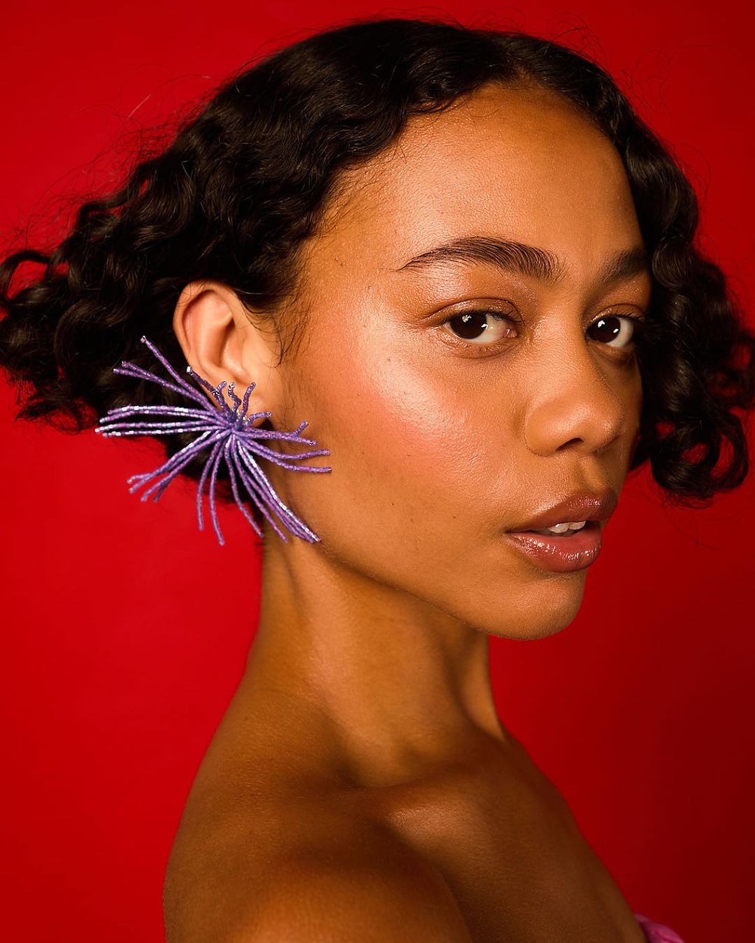 Close-up portrait of a woman with dark curly hair wearing purple earrings against a vibrant red background in a photo studio.