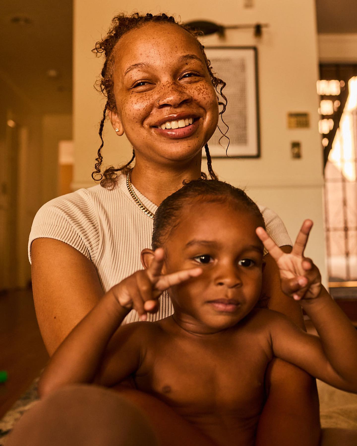Smiling mother and child pose indoors. The child gives a peace sign with their fingers, adding a playful touch to the warm scene.