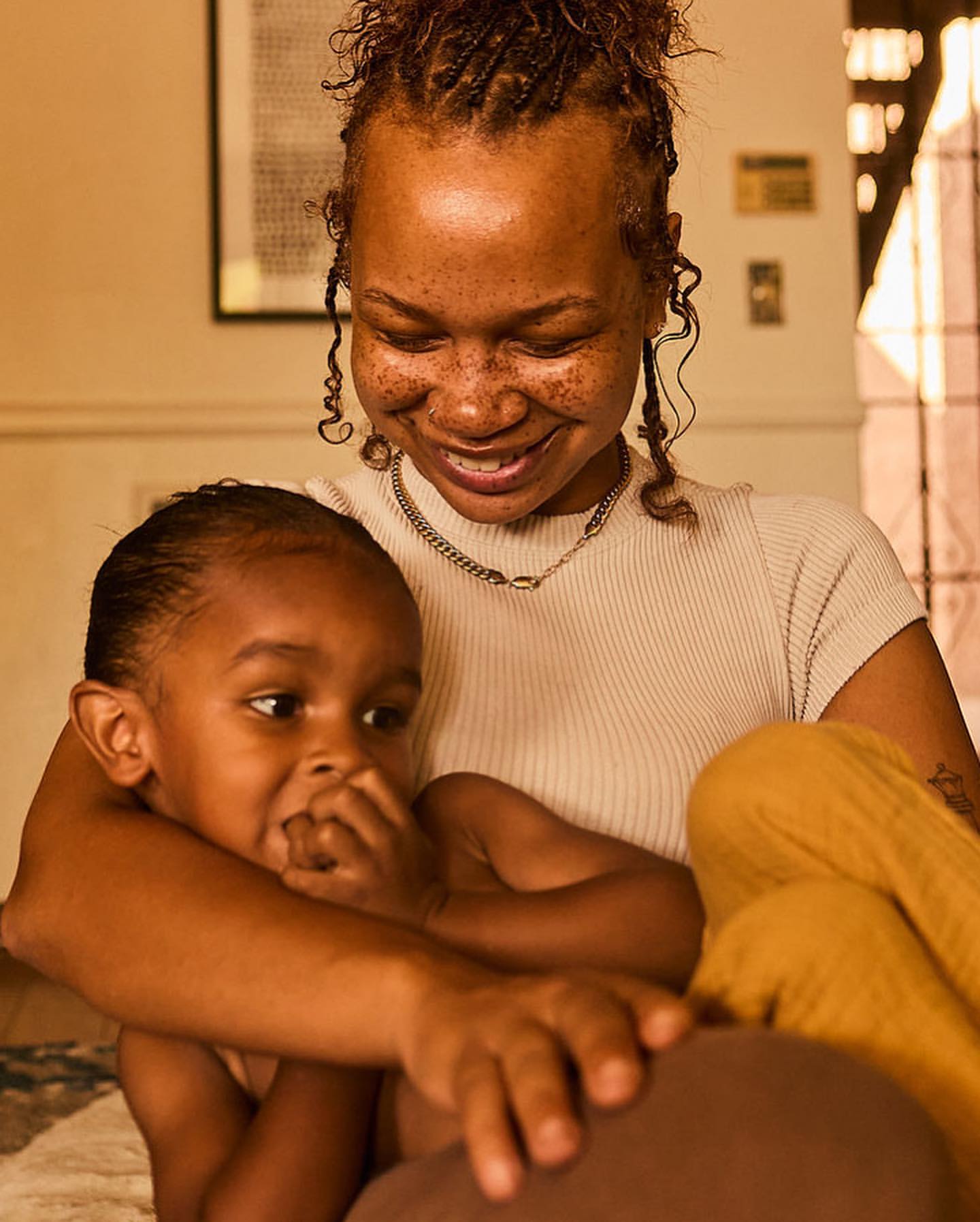 A smiling African-American mother embraces her young child, both sitting indoors. The child looks to the left, lit by warm light.
