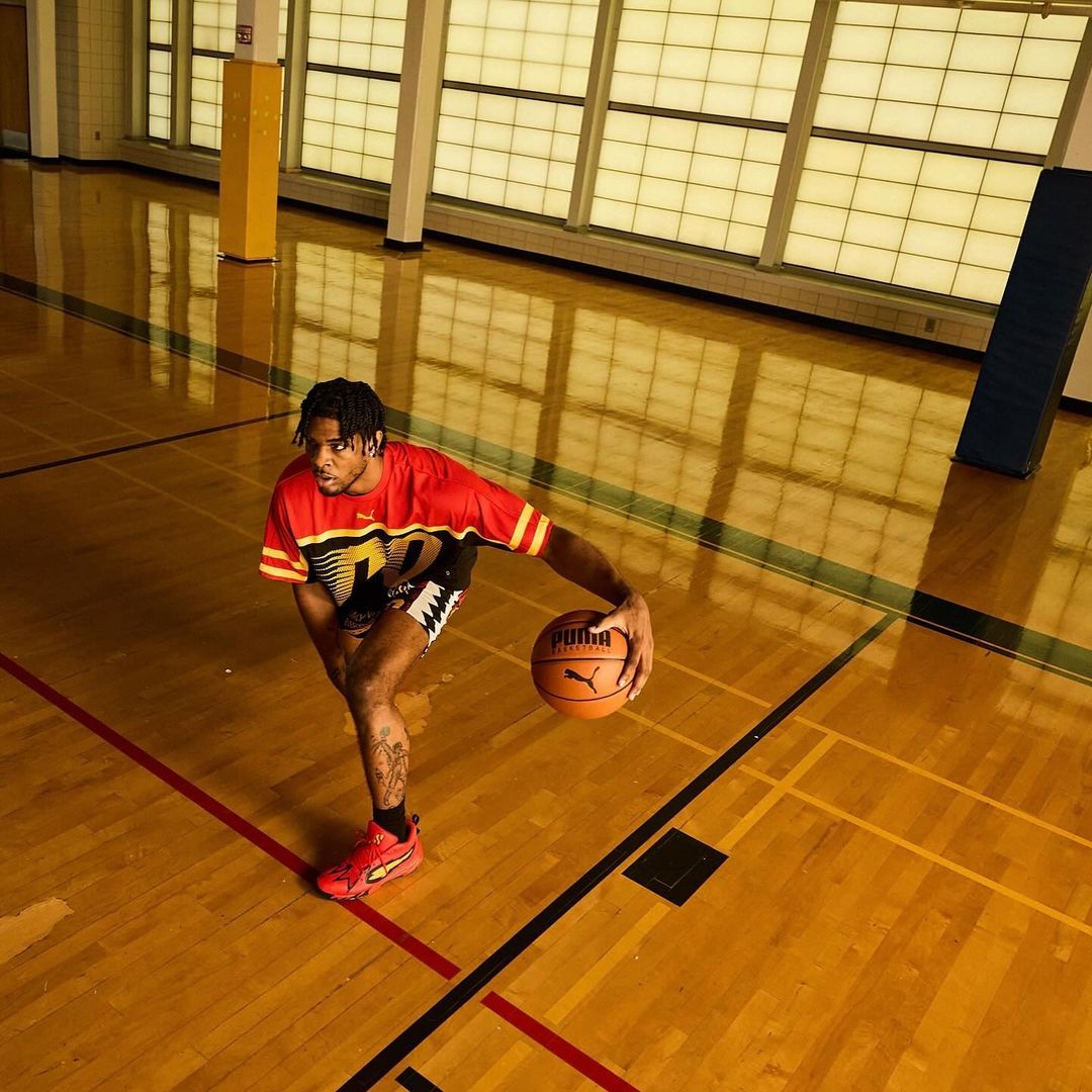 Male basketball player in a red Puma jersey dribbles a ball on a hardwood court indoors with natural light, wearing red Puma sneakers.