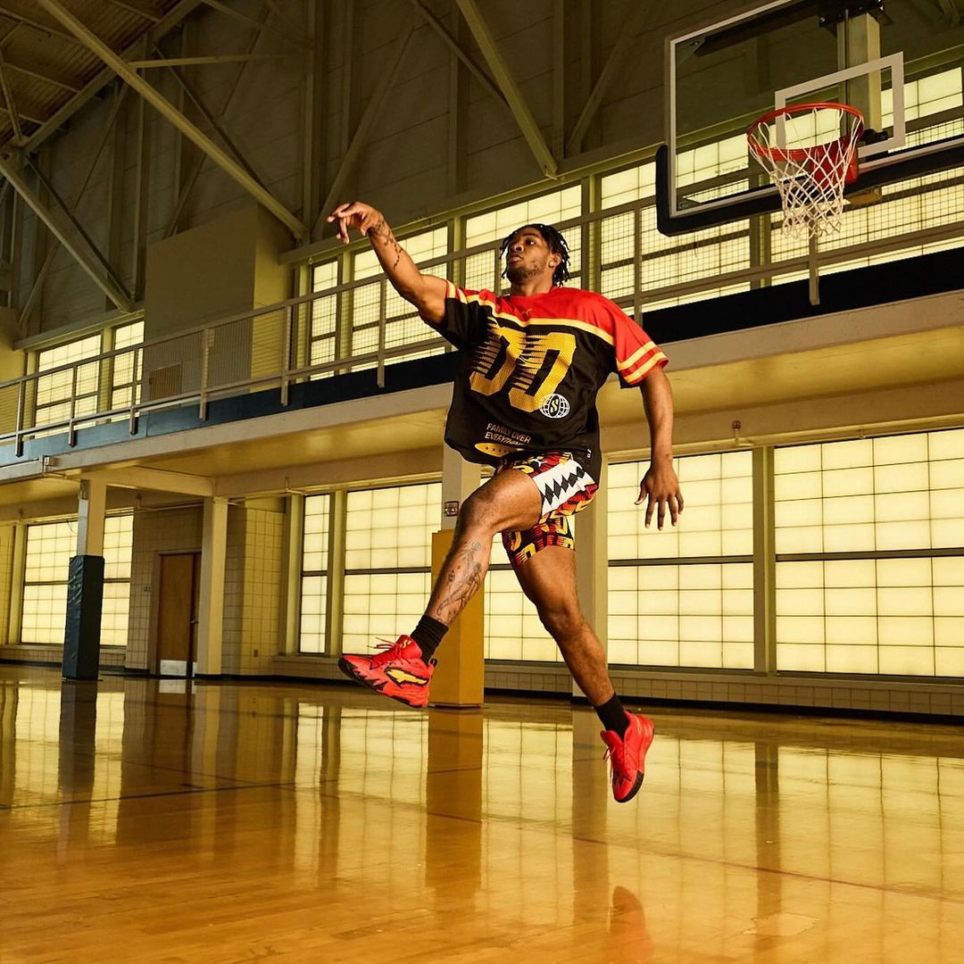 Athletic man in red sneakers jumps on basketball court, reaching up to hoop; indoor gymnasium has window and steel-truss roof.