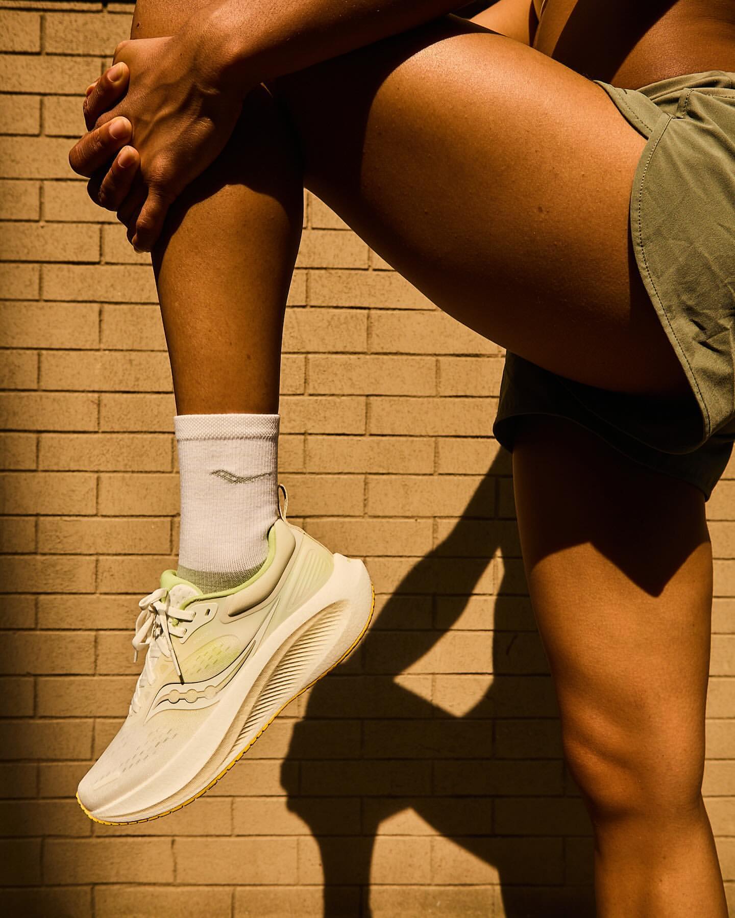 Close-up portrait of a runner wearing white running shoes, white socks, and green shorts against a brick wall.