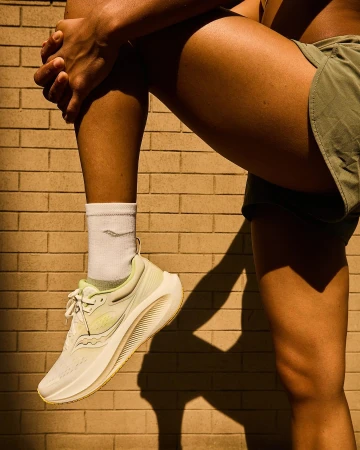 Close-up portrait of a runner wearing white running shoes, white socks, and green shorts against a brick wall.