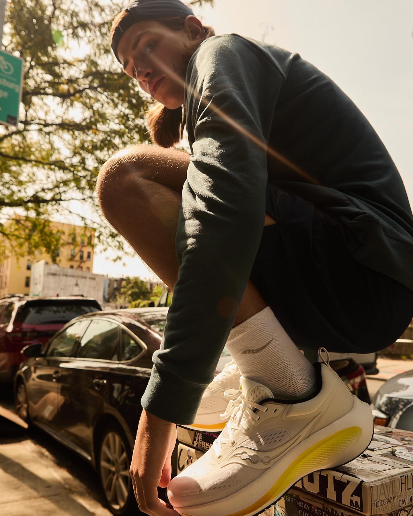 Man in athletic wear crouches beside a city street wearing Saucony running shoes with cars and buildings in the background on a sunny day.
