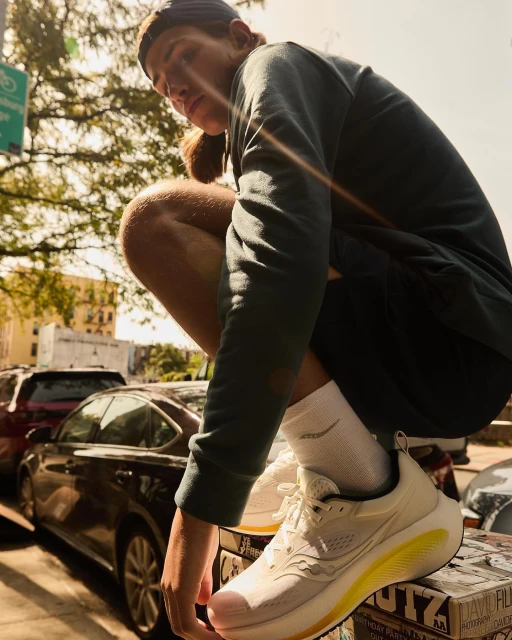 Man in athletic wear crouches beside a city street wearing Saucony running shoes with cars and buildings in the background on a sunny day.