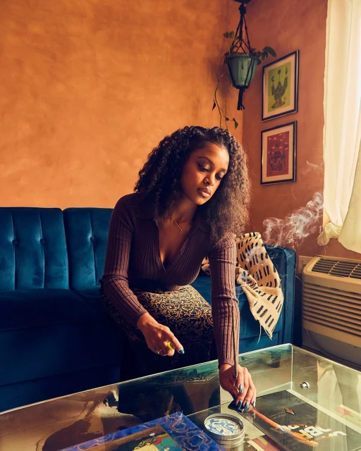 Young woman with curly hair sits indoors with a glass coffee table with art magazines and incense stick burning in a cozy living room.