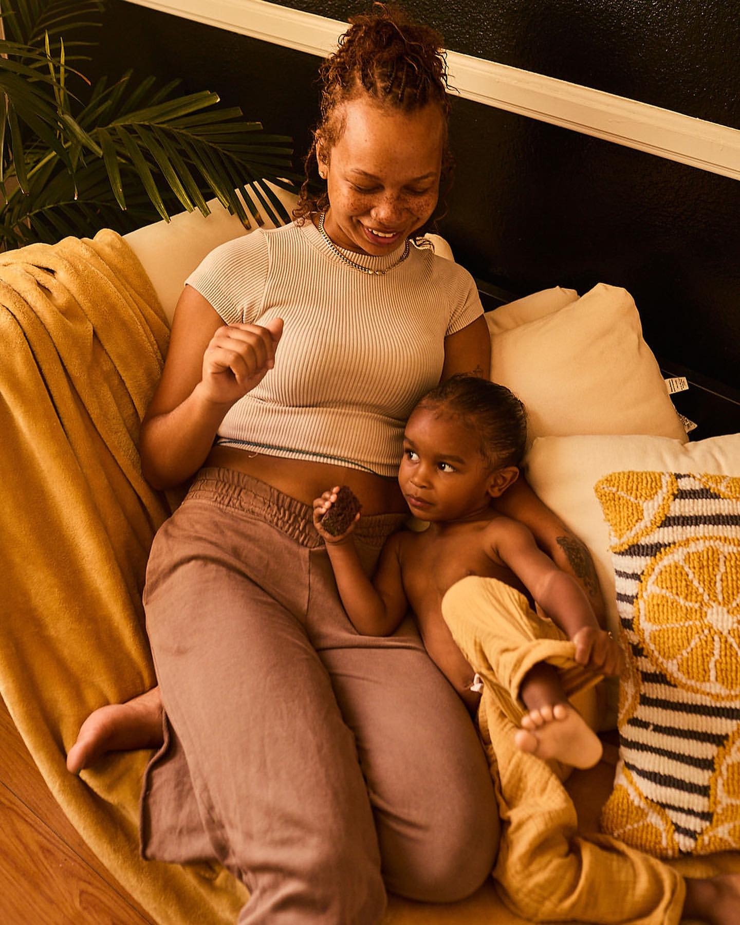 Mother and child relaxing together on a sofa. A woman is holding a snack, while a young child sits beside her in a comfortable indoor setting.