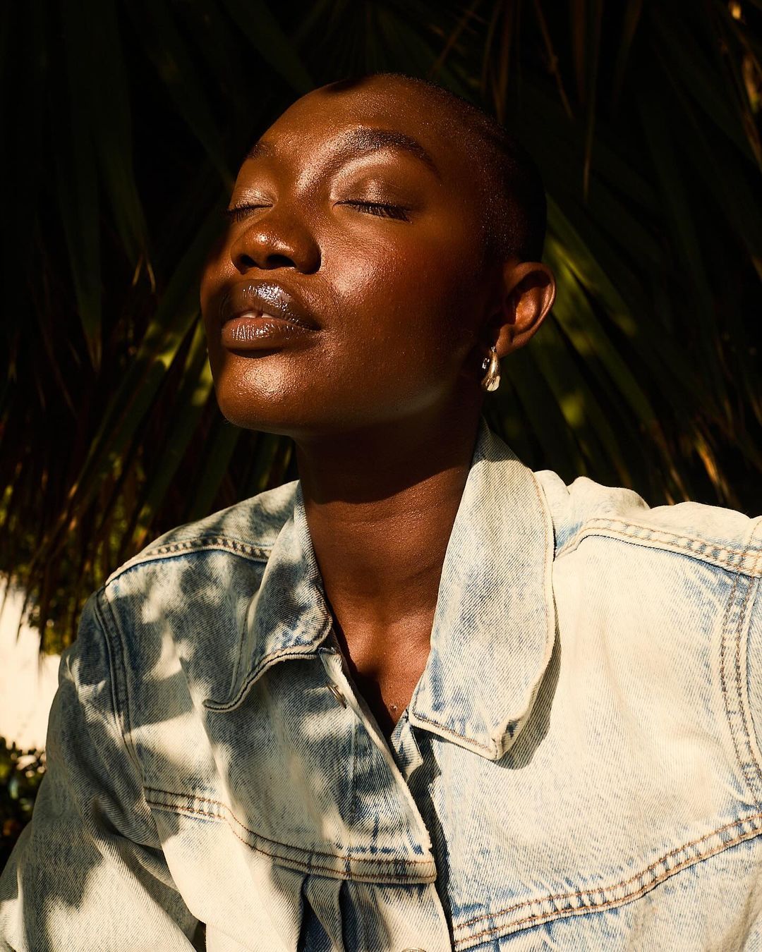 Close-up of a Black woman with eyes closed, wearing a denim jacket, basking in sunlight against a backdrop of green palm fronds.
