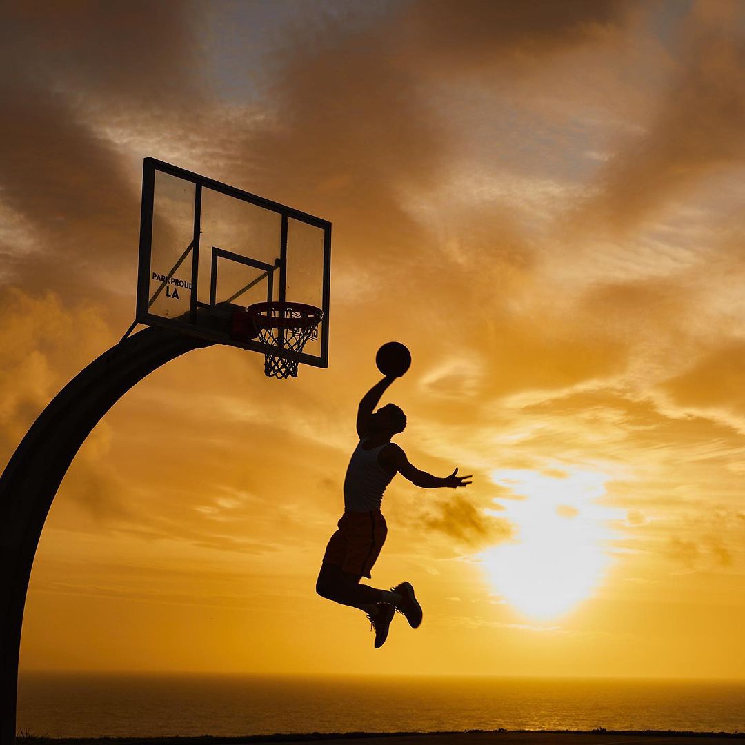 Silhouette of adult male basketball player in mid-air leap shooting a basketball at a hoop against a sunset and ocean backdrop.