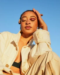 Close-up portrait of a woman with freckles, braided hair beads, red eyeshadow, and cream utility jacket against a clear blue sky