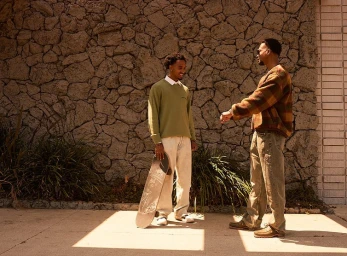 Two Black men standing outdoors holding a skateboard against a textured stone wall background