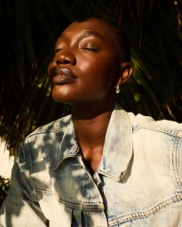 Close-up portrait of a bald Black woman wearing a light denim jacket outdoors with dappled sunlight on her face.