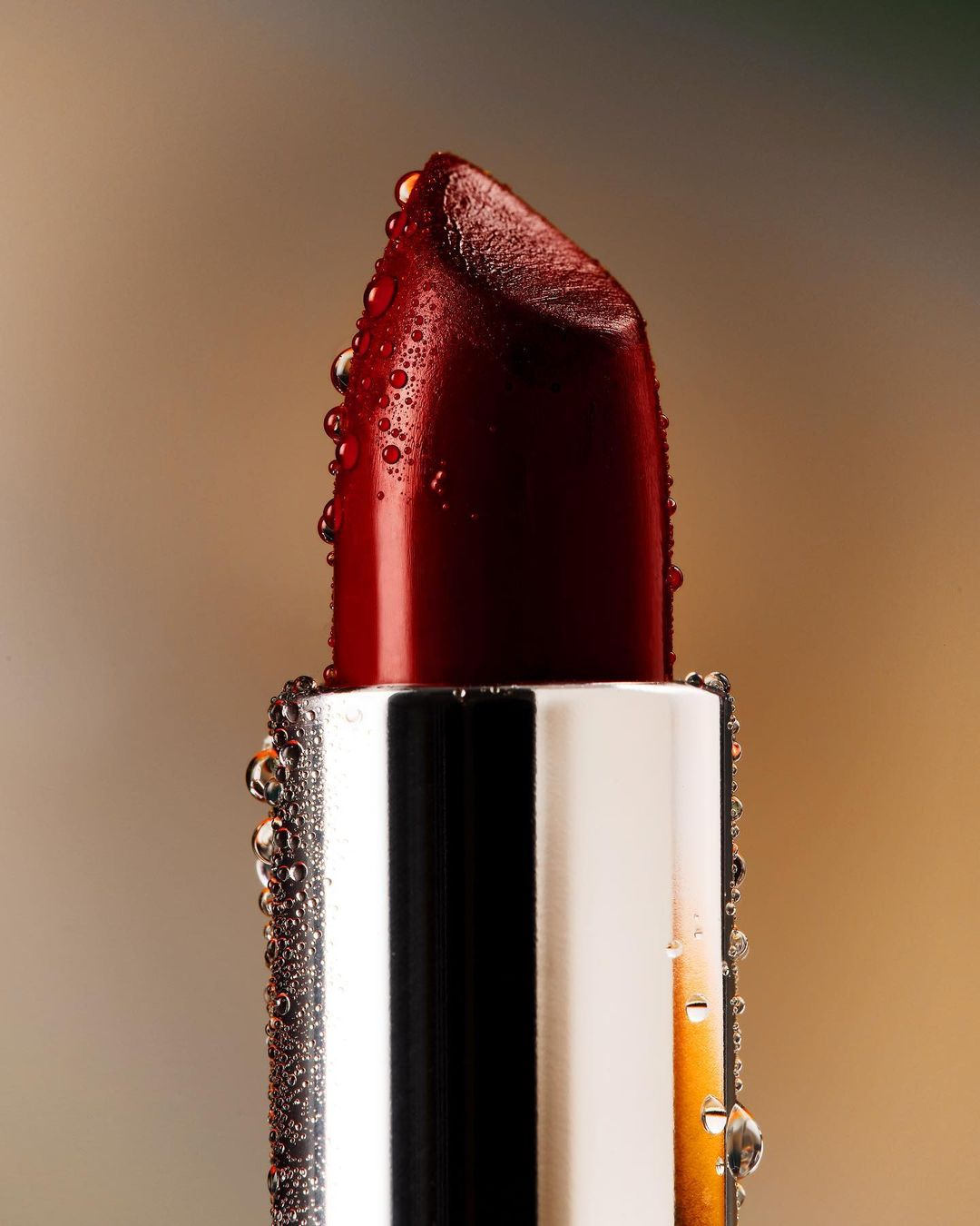Macro shot of a dark red lipstick bullet with water droplets on a silver tube against a blurred brown and gold background.