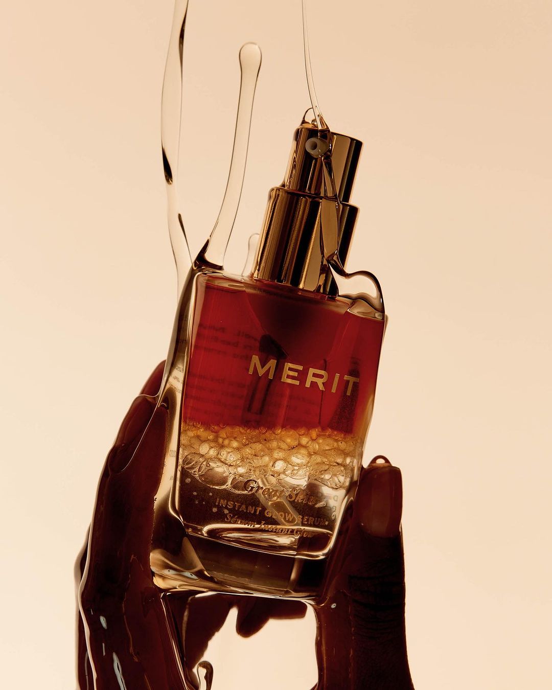 Cosmetic product by Merit held in a person's hand with a translucent liquid pouring over it. The product is reddish-brown.