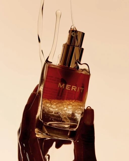 Cosmetic product by Merit held in a person's hand with a translucent liquid pouring over it. The product is reddish-brown.