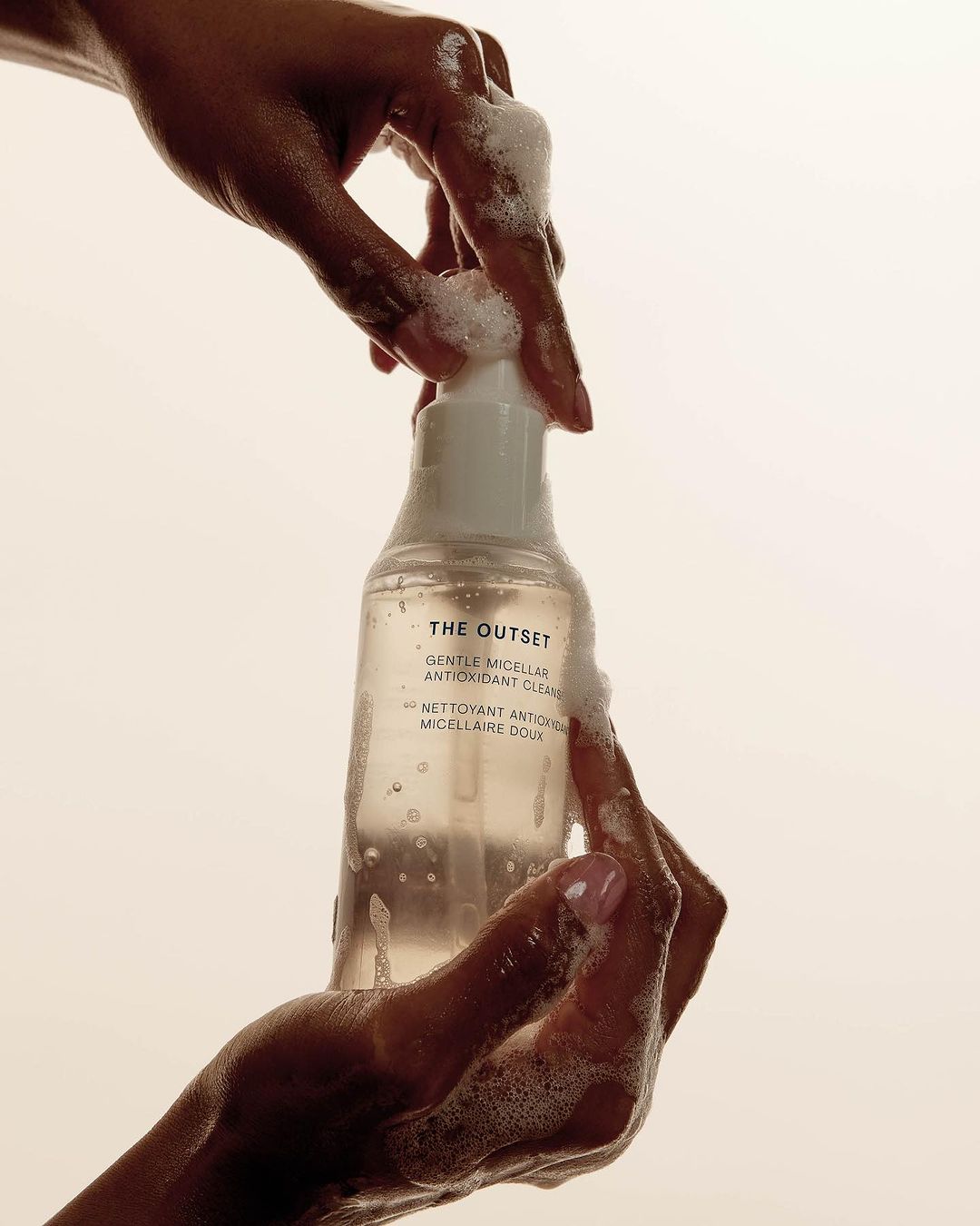Close-up of hands holding a bottle of 'The Outset' antioxidant cleanser. Soap bubbles and neutral studio background.