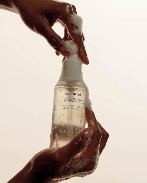 Close-up of hands holding a bottle of 'The Outset' antioxidant cleanser. Soap bubbles and neutral studio background.