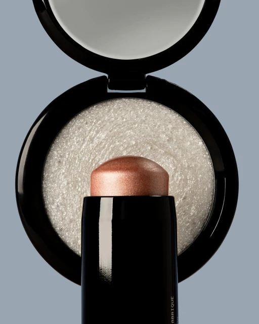 Close-up studio shot of a black and rose gold highlighter stick with a reflective compact against a gray background, product photography.
