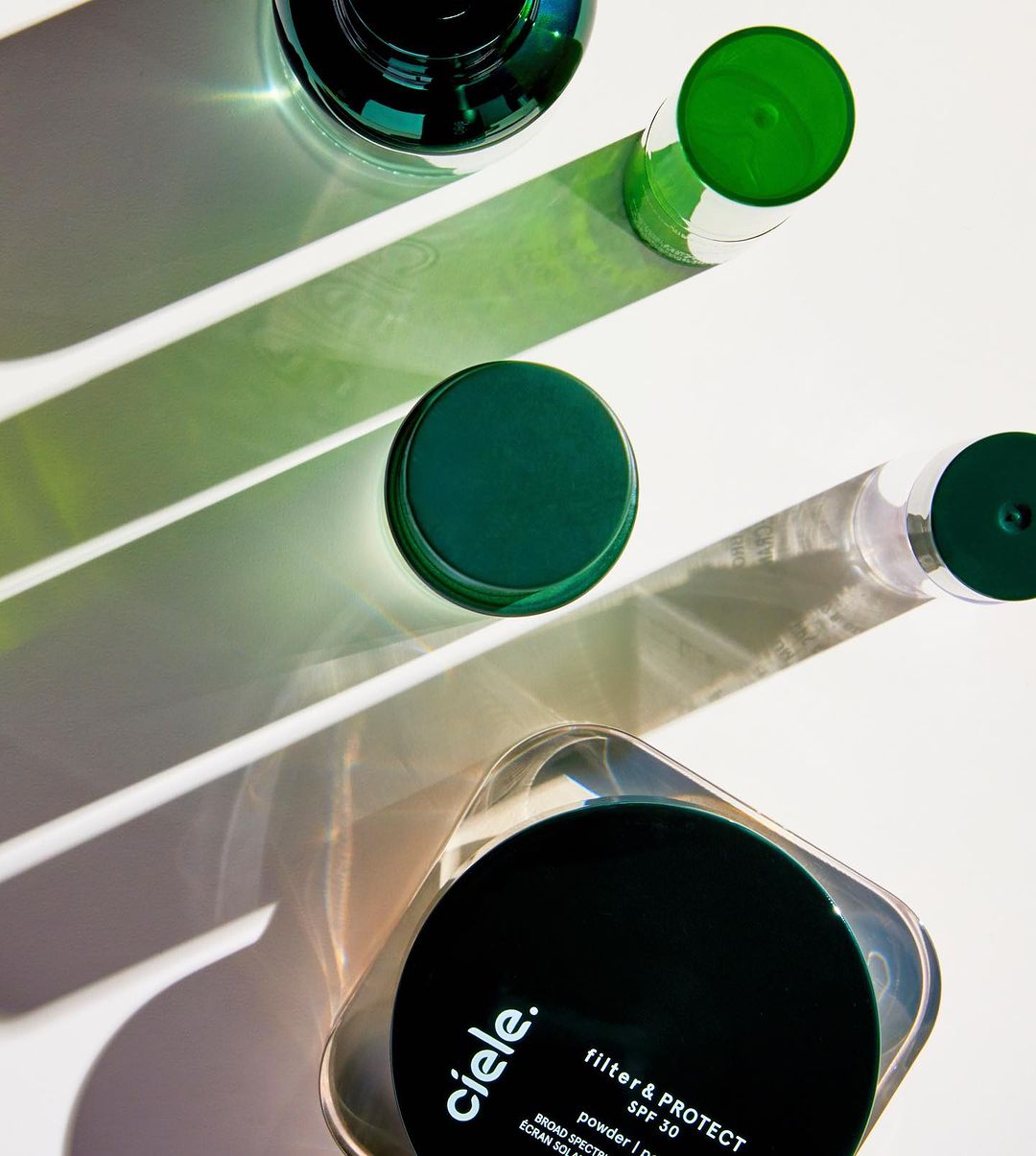 Overhead shot of Ciele cosmetics containers and bottles in varying sizes, with light and shadow play on a solid white surface.