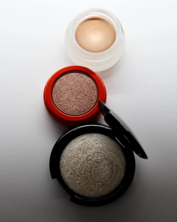 Overhead view of makeup: three cosmetic containers, including eyeshadow and highlighter in neutral tones and sleek black eyeliner.