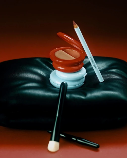 Makeup still life features powder compacts, makeup brush, and eyeliner pencil on a black cushion with a warm gradient background.