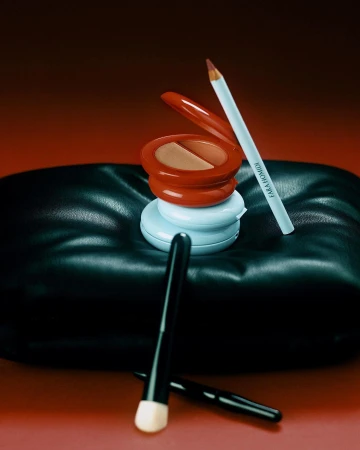 Makeup still life features powder compacts, makeup brush, and eyeliner pencil on a black cushion with a warm gradient background.