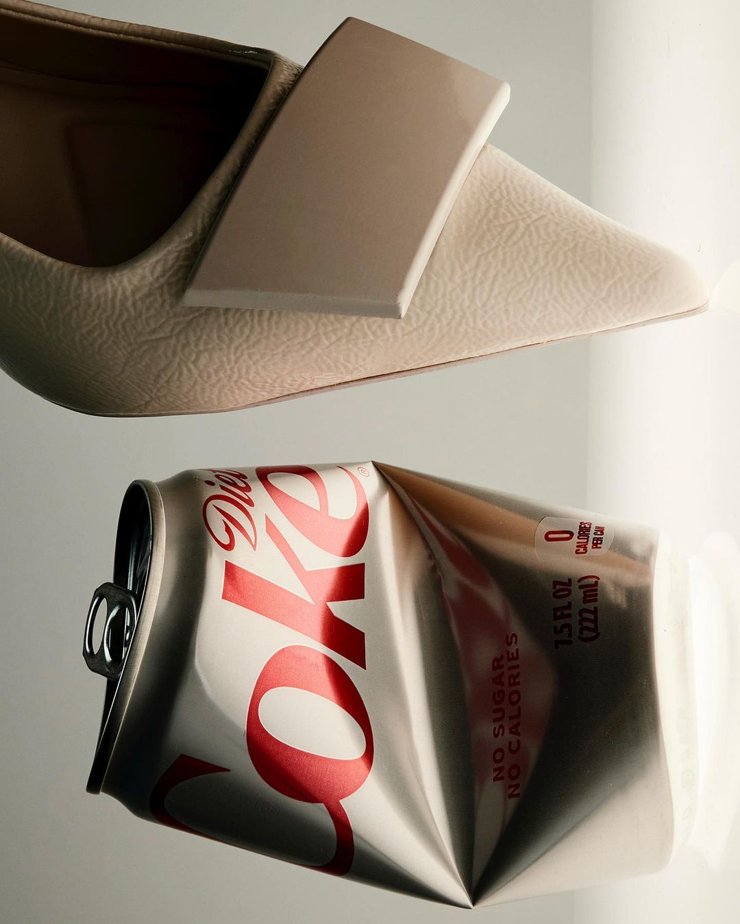 Still life of a beige leather pump shoe beside a crushed Diet Coke can. Shot in a photo studio with neutral colors and soft light.