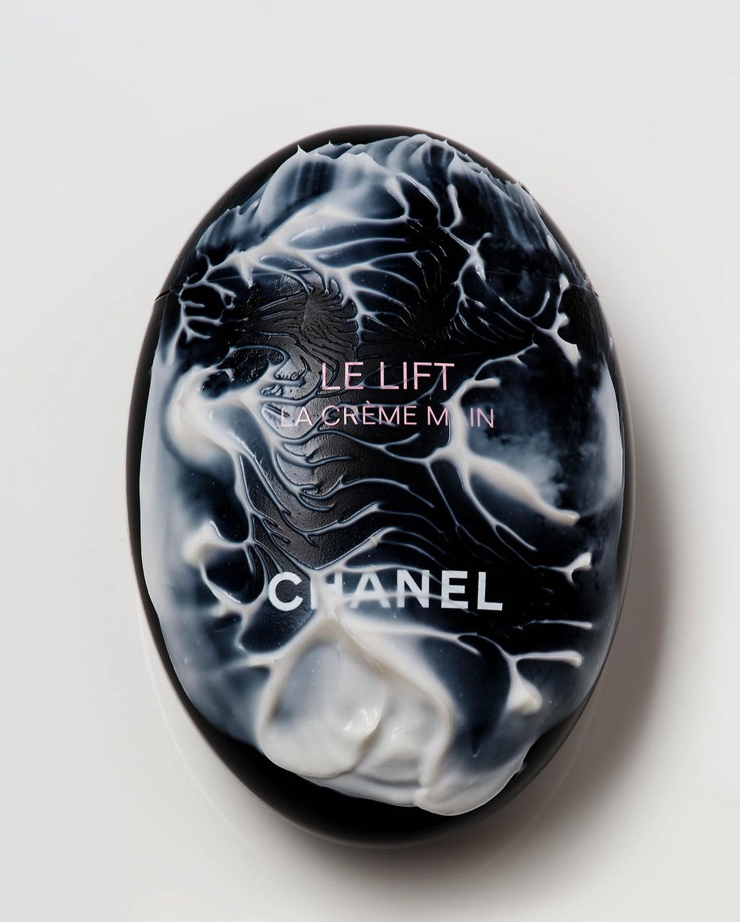 Close-up of Chanel Le Lift La Crème Main hand cream in an egg-shaped black case with white cream accents on a white background.