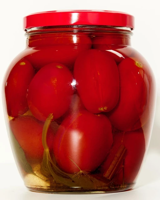 A glass jar filled with red pickled tomatoes and a red lid stands out against a bright white background, showcasing preserved food.
