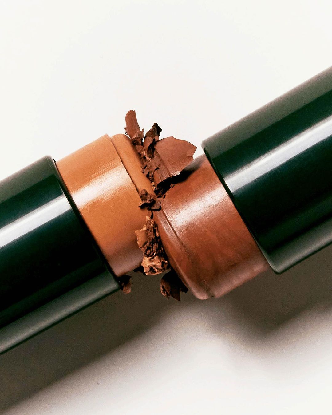 Broken brown makeup stick with a dark green tube on a white surface. Cosmetic product, close-up shot, studio lighting, beauty concept.