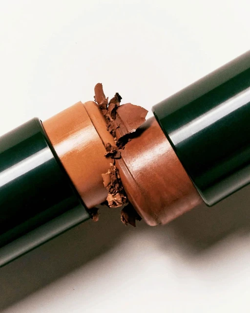 Broken brown makeup stick with a dark green tube on a white surface. Cosmetic product, close-up shot, studio lighting, beauty concept.