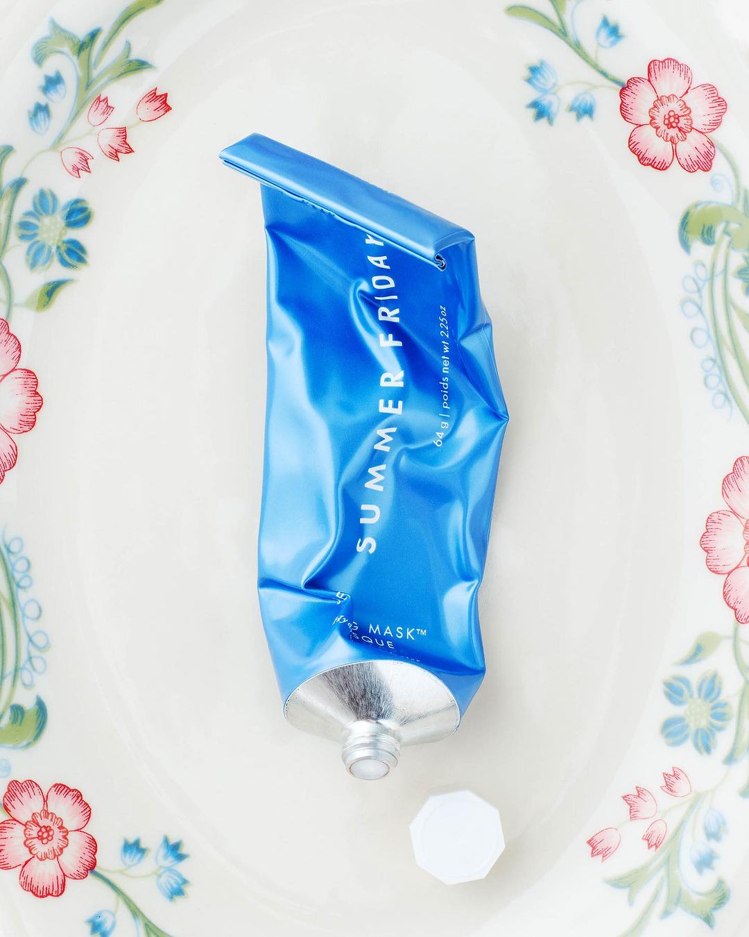 Summer Friday's blue Jet Lag Mask tube with its white cap on a white plate decorated with floral design, top-down studio shot.