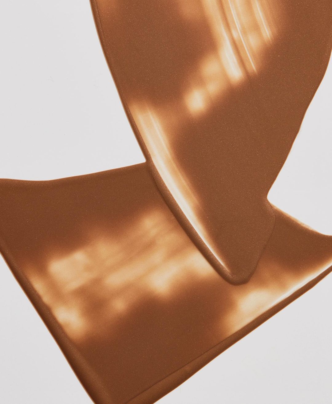 Close-up studio shot of a swirl of light brown foundation makeup, revealing a smooth, creamy texture against a clean white backdrop.