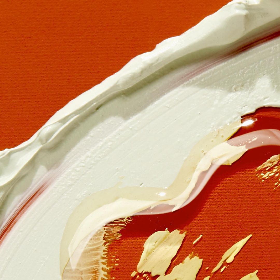 Macro shot of abstract painting with orange canvas background and a thick, curving application of white and gold paint.