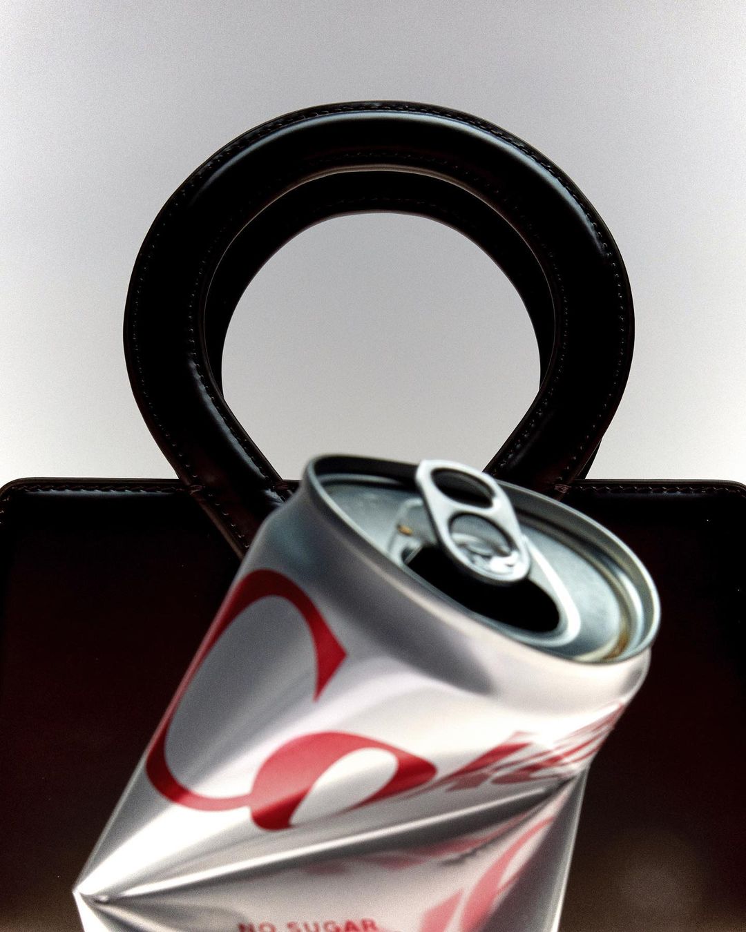A crumpled Coca-Cola can sits in front of a dark handbag with a round handle against a plain white backdrop in a studio shot.