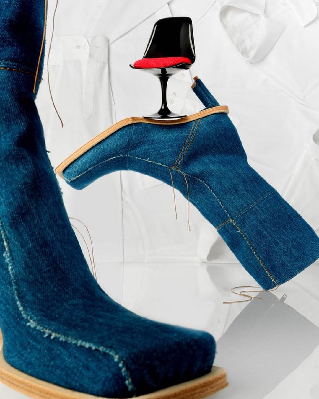 Denim heeled boots with a miniature red-seat chair and white collared shirts create a surreal fashion composition on a white surface.