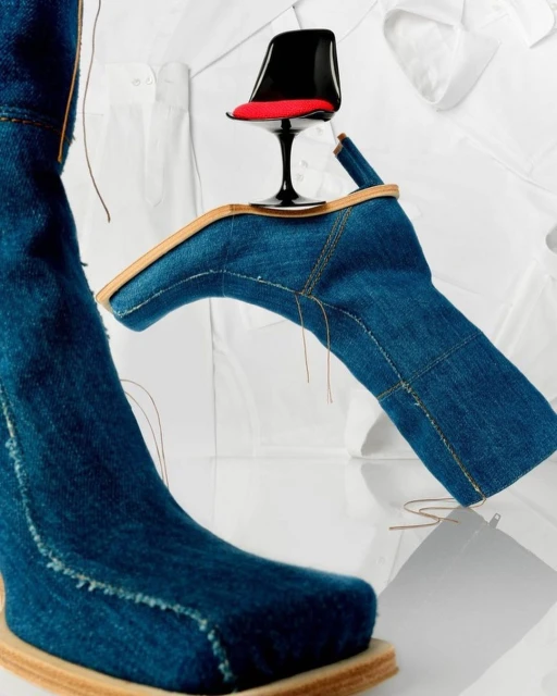 Denim heeled boots with a miniature red-seat chair and white collared shirts create a surreal fashion composition on a white surface.