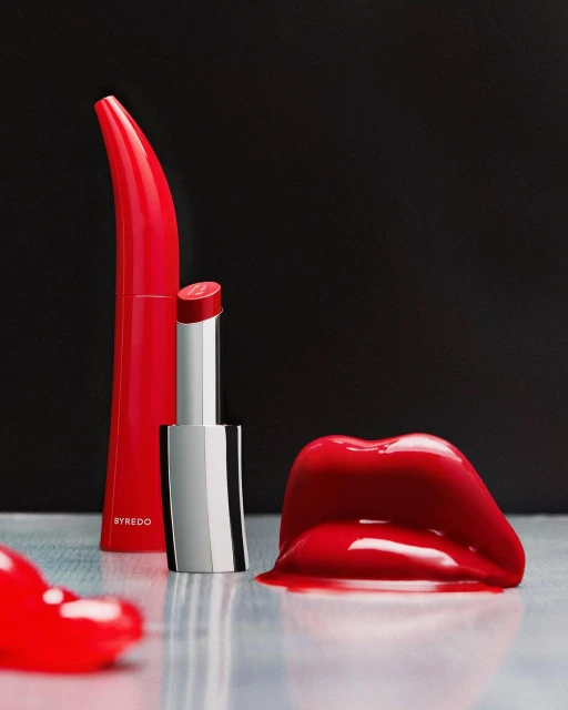 Red lipstick tubes and a glossy lip-shaped object are arranged on a reflective surface against a dark background in a studio shot.