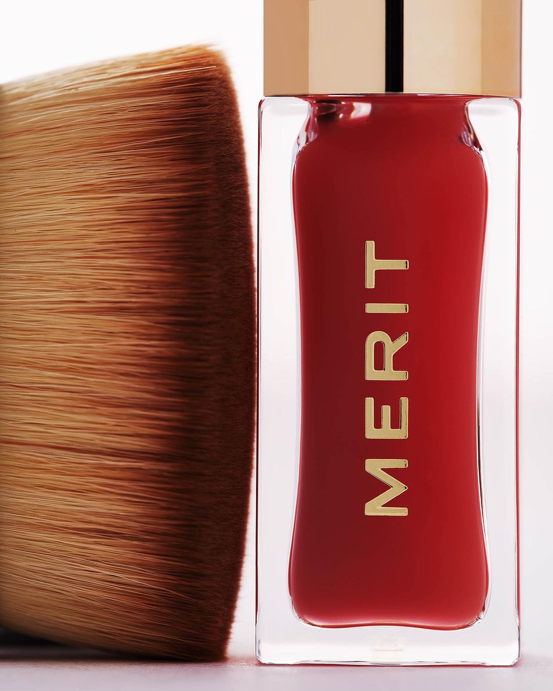 Close-up of a makeup brush and Merit beauty product in a clear container with red liquid, highlighted on a white background.