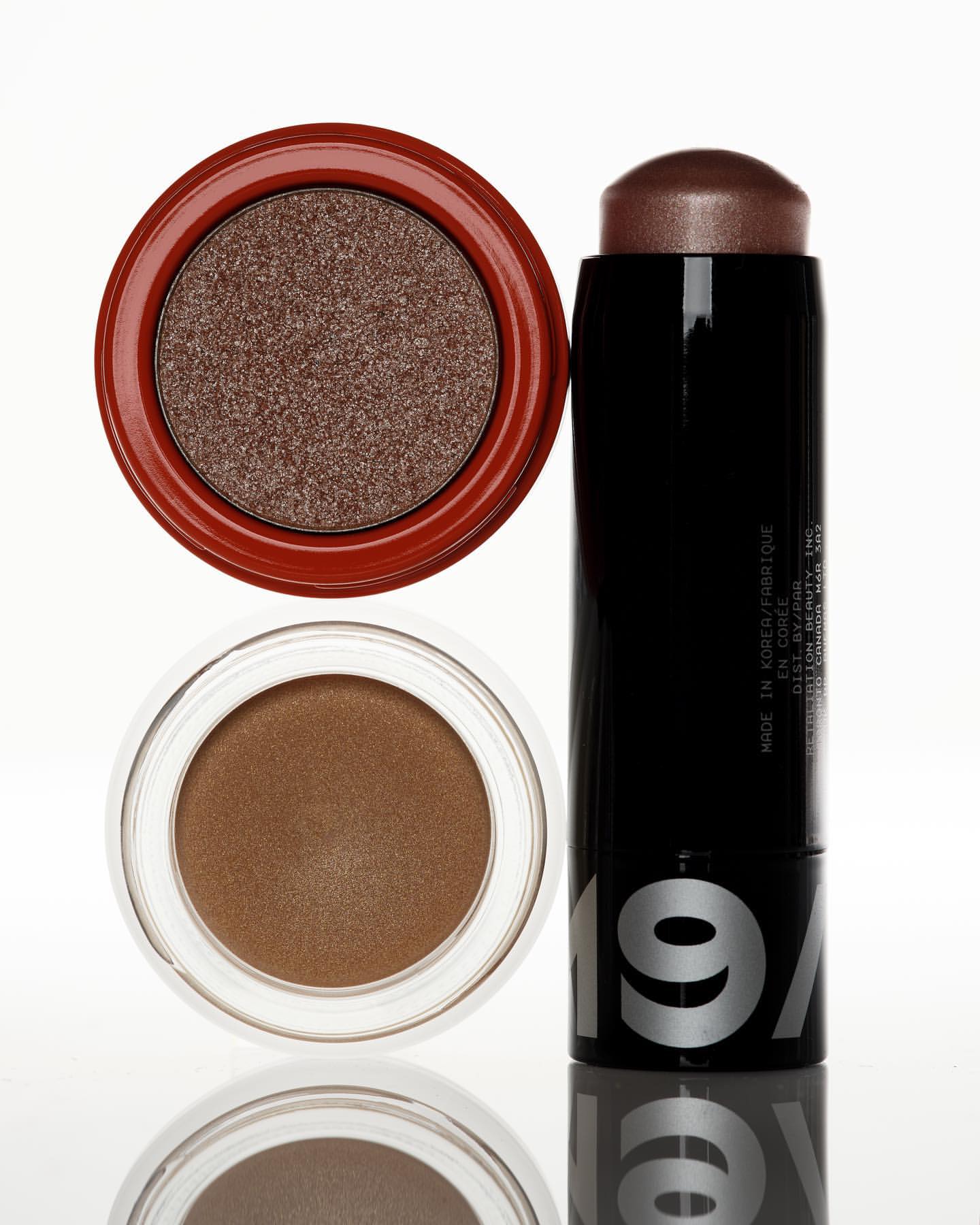 Still life of makeup with two eyeshadow compacts, one with reddish brown shadow and another with bronze shadow, and a black makeup stick.