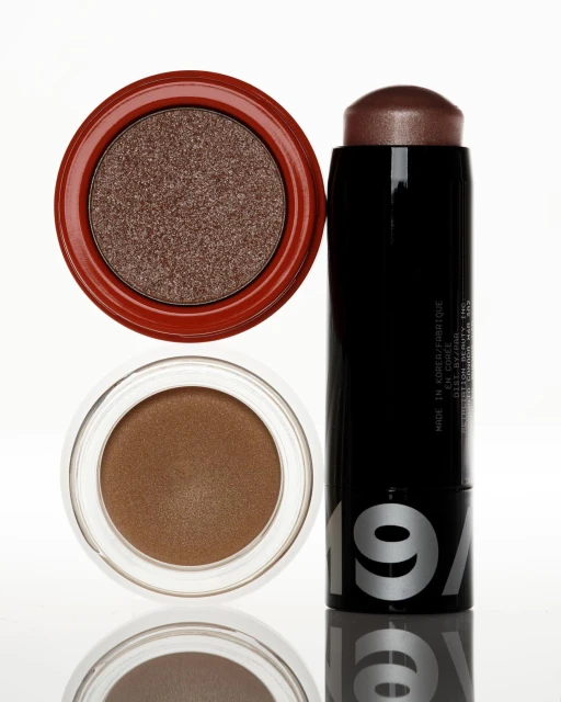 Still life of makeup with two eyeshadow compacts, one with reddish brown shadow and another with bronze shadow, and a black makeup stick.