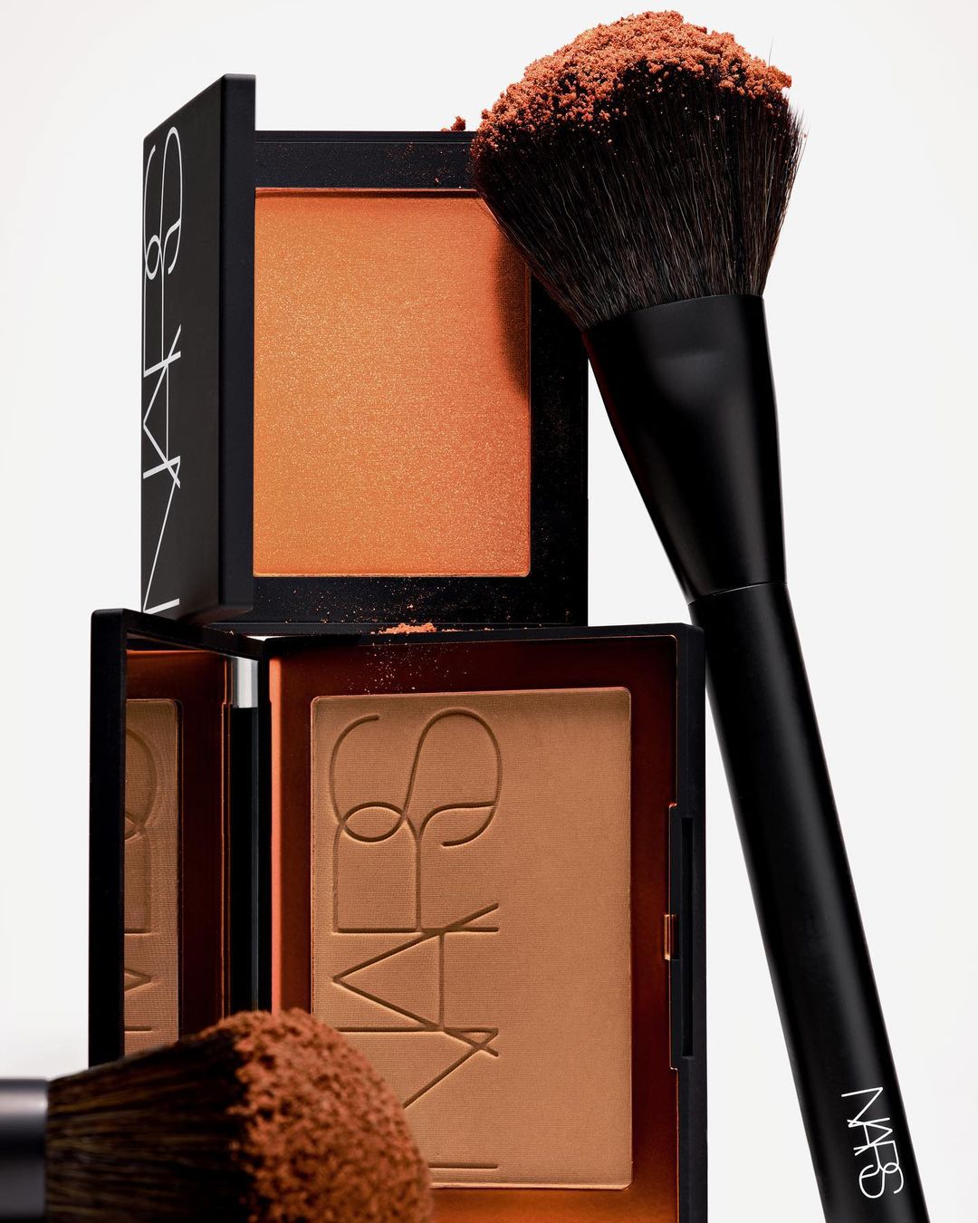 Makeup still life featuring NARS bronzer and blush compacts stacked together with a black makeup brush against a white backdrop.