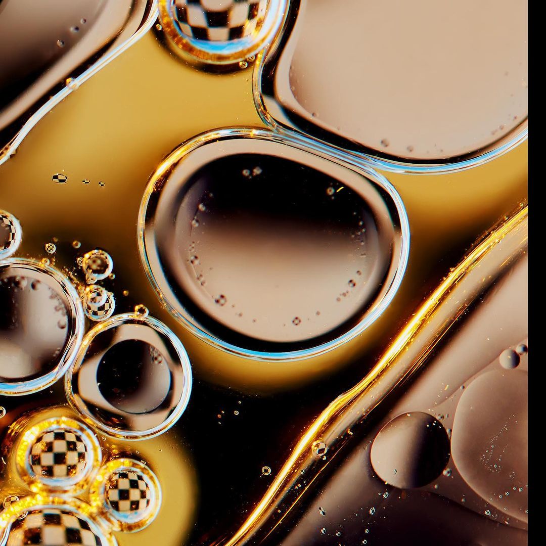 Abstract macro photograph of oil and water mix with iridescent bubbles, refracted checkered pattern, and a shiny gold background.