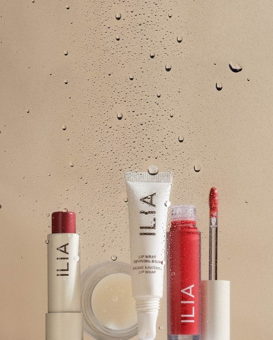 Close-up studio shot of Ilia brand lip care products including balm, lipstick, and lip gloss displayed against a water droplet background.