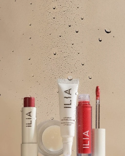 Close-up studio shot of Ilia brand lip care products including balm, lipstick, and lip gloss displayed against a water droplet background.