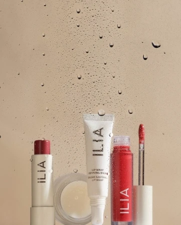 Close-up studio shot of Ilia brand lip care products including balm, lipstick, and lip gloss displayed against a water droplet background.
